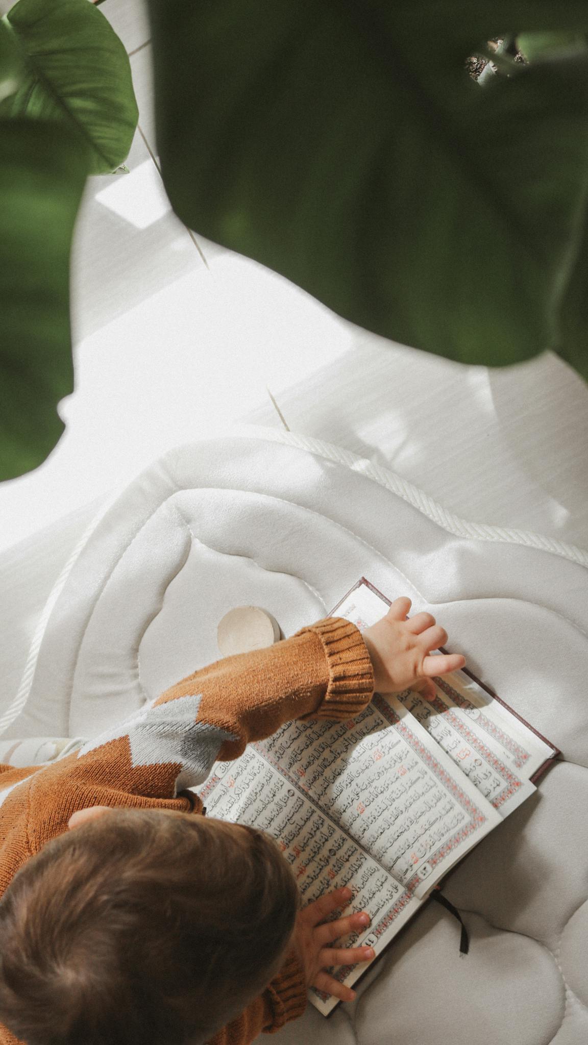 Overhead view of a child reading the Quran with focus on hands, highlighting a moment of learning and spirituality.