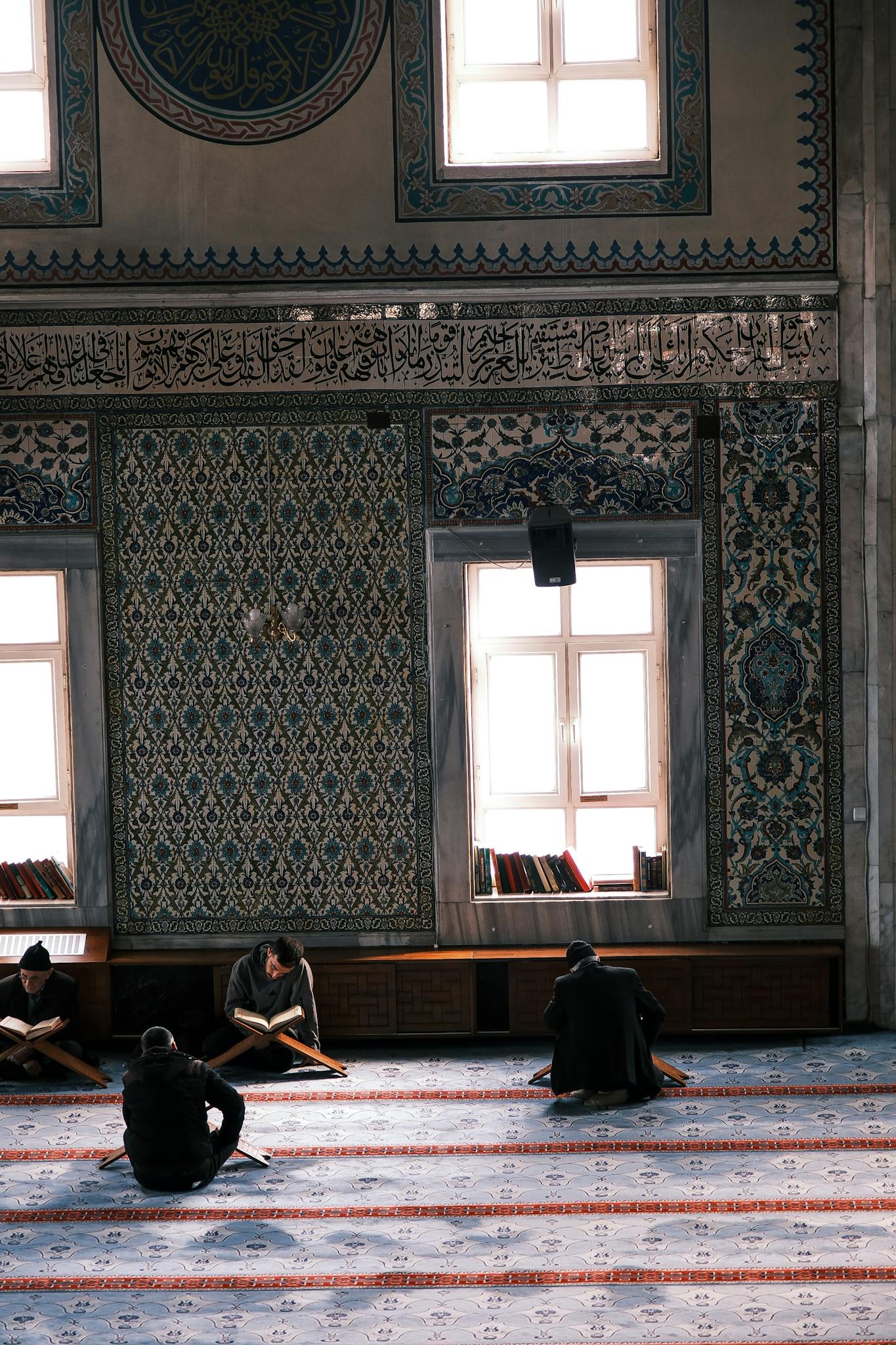 People reading in a beautifully decorated mosque interior with intricate patterns and sunlight.
