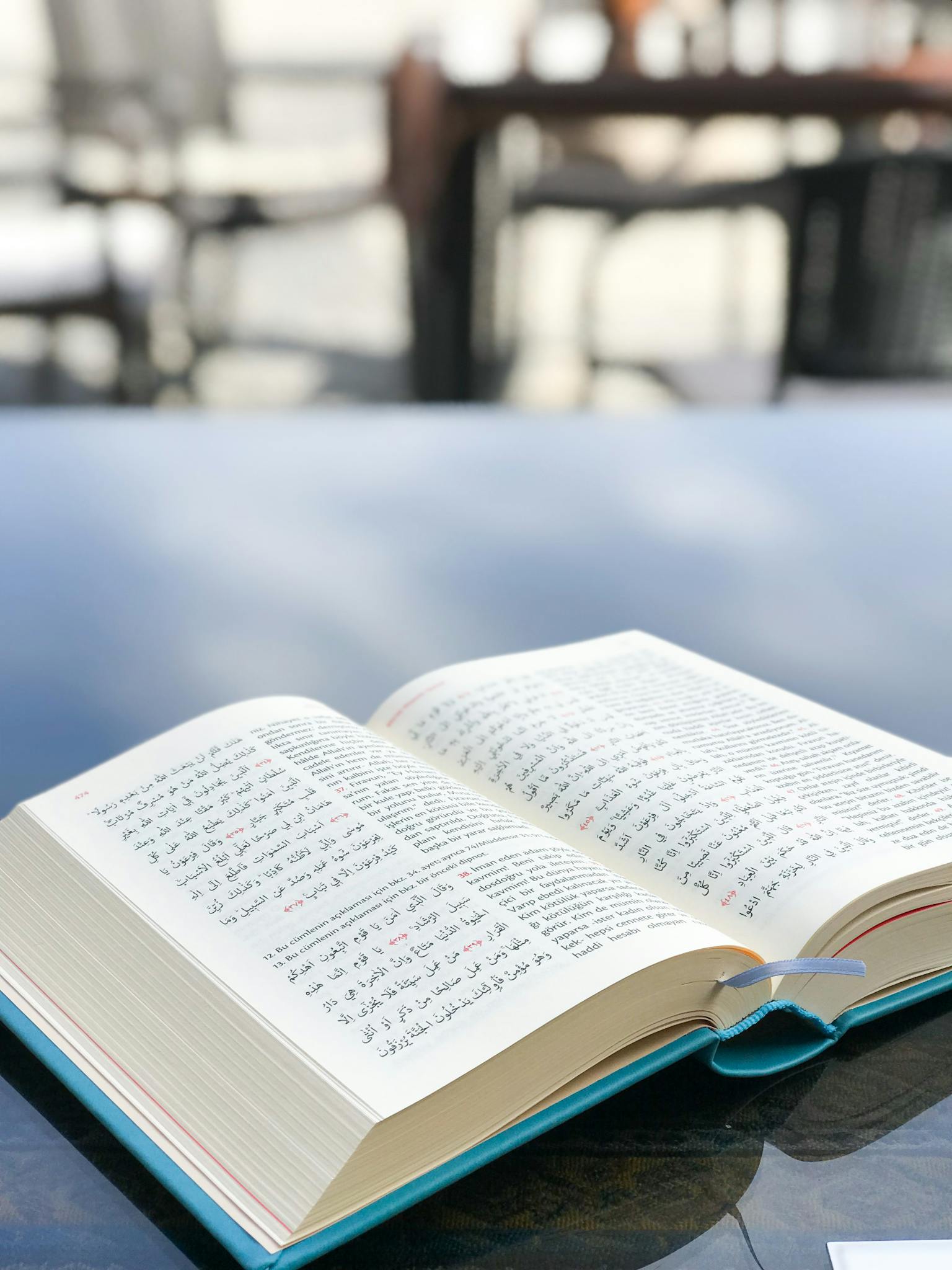 An open Arabic book rests on a glass table with blurred outdoor background.